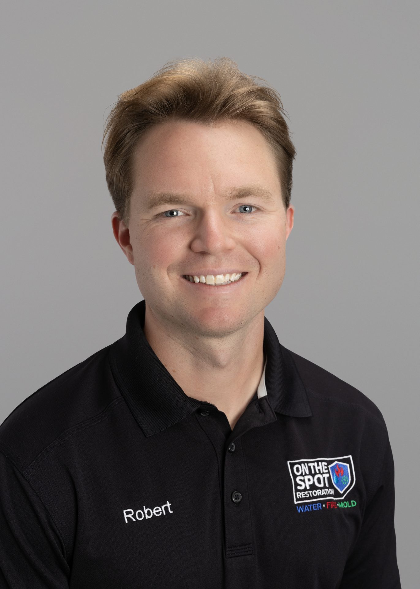 A smiling man with light brown hair wearing a black polo shirt, featuring a name tag 'Robert' and a logo on the chest.