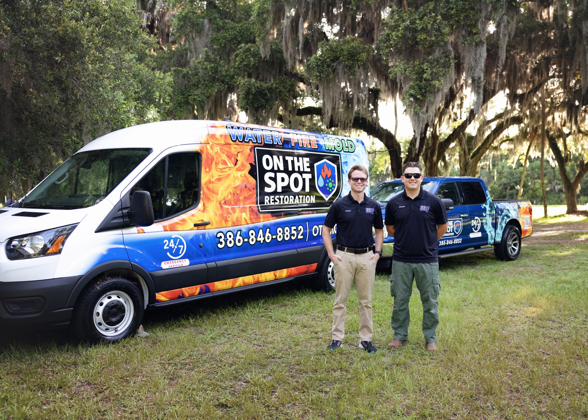 Two individuals stand beside a branded service van and pickup truck, surrounded by trees and greenery.