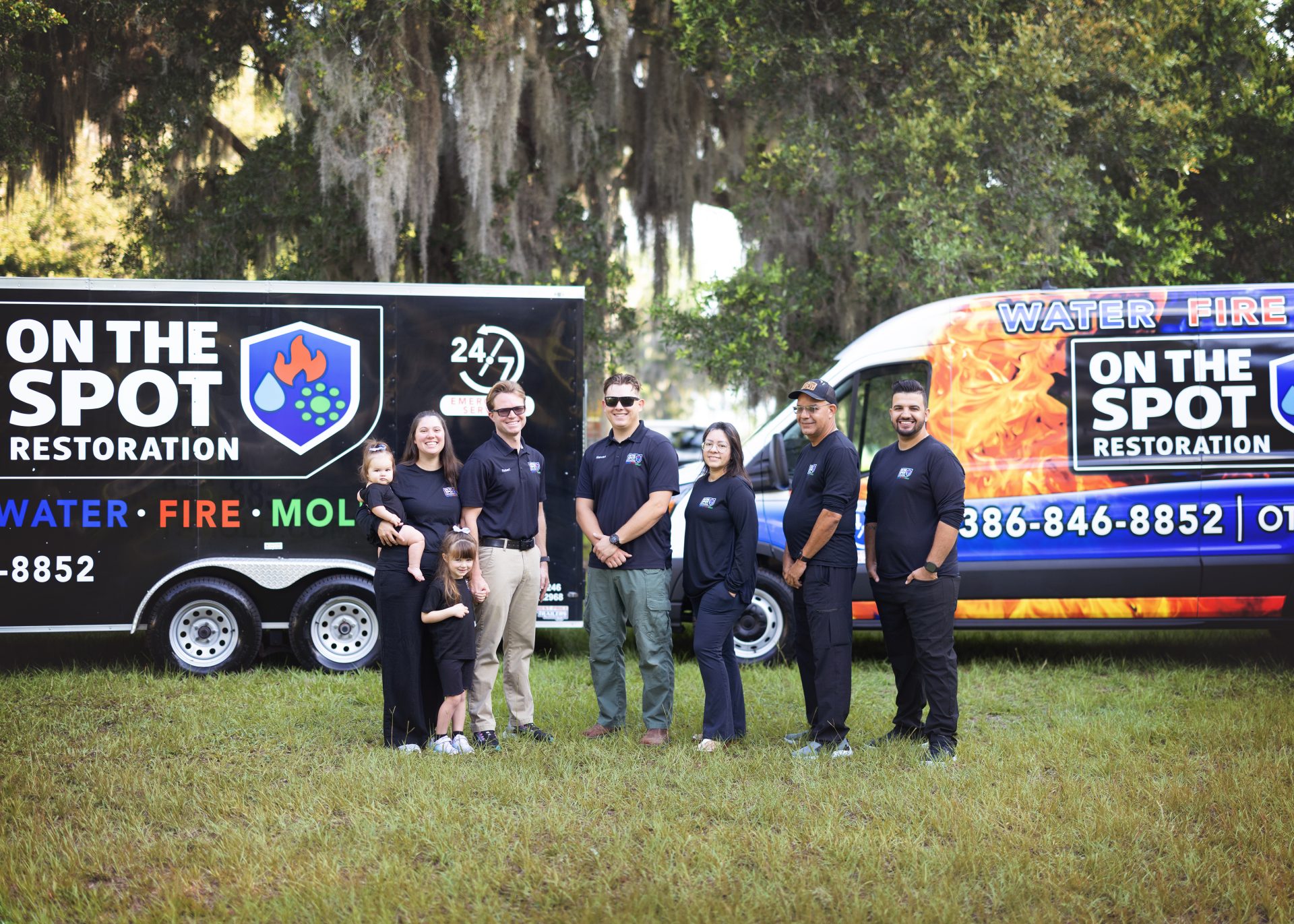 A group of six people in navy shirts stand in front of branded vehicles for a restoration service in a grassy area.