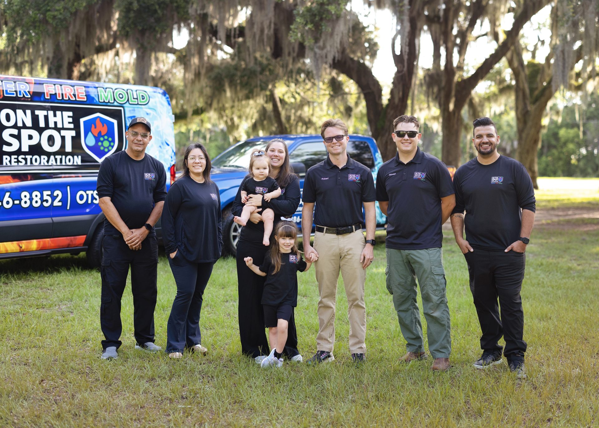 A group of people in black shirts standing in a grassy area, with a branded vehicle in the background and trees nearby.