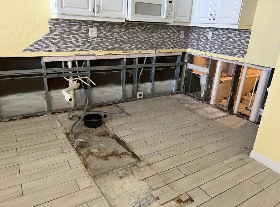 A partially renovated kitchen with exposed plumbing and flooring, showcasing a combination of tiled and removed sections. | On The Spot Restoration