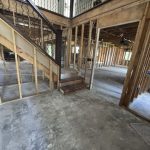 An interior view of a partially constructed home, showcasing exposed wooden framing, an unfinished staircase, and an open floor layout.