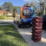 A stacked pile of equipment sits on a driveway near a brightly wrapped van labeled 'ON THE SPOT RESTORATION' in a residential area.