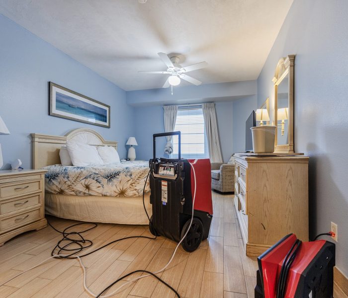 A cozy bedroom with blue walls, featuring a bed, nightstands, lamps, a television, and bags on the floor.