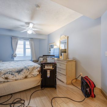 A cozy bedroom with a blue theme, featuring a bed, dresser, and a fan, accompanied by some equipment and cords on the floor.