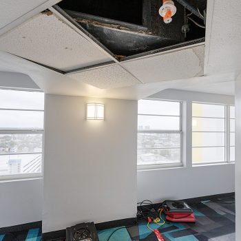 An unfinished interior space with a removed ceiling tile, visible pipes, and drying equipment on a patterned carpet.