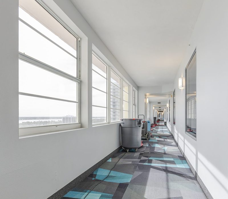 A long, brightly lit corridor featuring patterned carpet, windows along one side, and cleaning equipment at the far end.