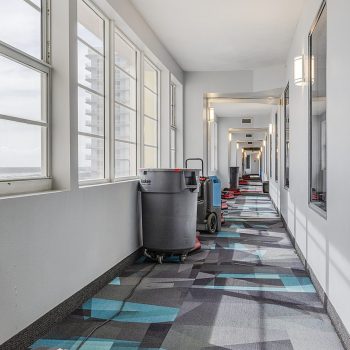 A modern hallway features colorful geometric carpets and cleaning equipment, illuminated by natural light through large windows.