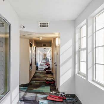 A long hallway with a patterned floor, bright windows, and several red vacuum cleaners stationed on the ground.