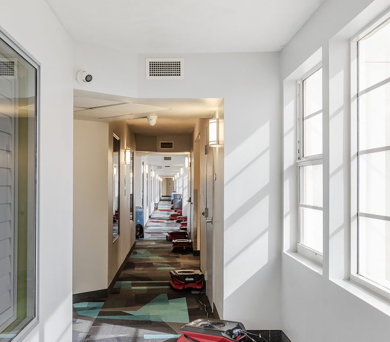 A long hallway with a patterned floor, bright windows, and several red vacuum cleaners stationed on the ground.