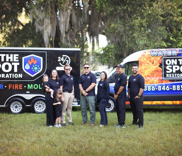 A group of six people in navy shirts stand in front of branded vehicles for a restoration service in a grassy area.